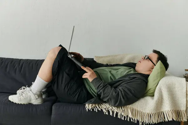 A man with Down syndrome relaxes on the couch, engaged with his laptop. — Stock Photo