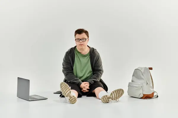 A young man with Down syndrome sits comfortably at home, engaged in quiet reflection with his laptop nearby. — Stock Photo