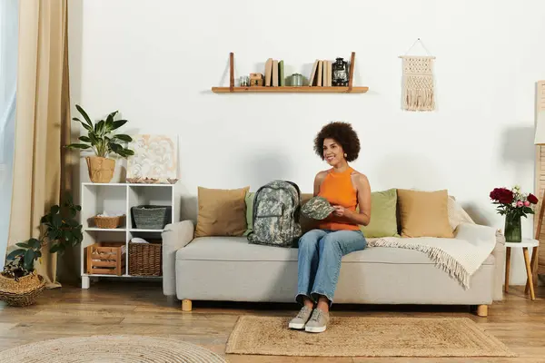 A young woman smiles while holding a camouflage backpack in her inviting living room. — Stock Photo
