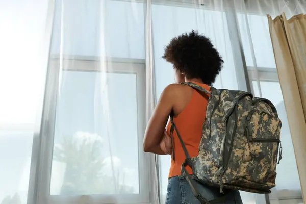 A young woman gazes thoughtfully out the window, her camouflage backpack resting on her back. — Stock Photo