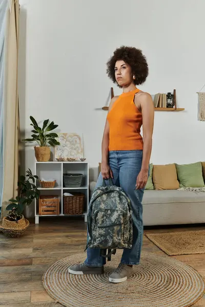 Una joven mujer está de pie con confianza en su sala de estar, luciendo una mochila de camuflaje y una vibrante camiseta naranja. — Stock Photo
