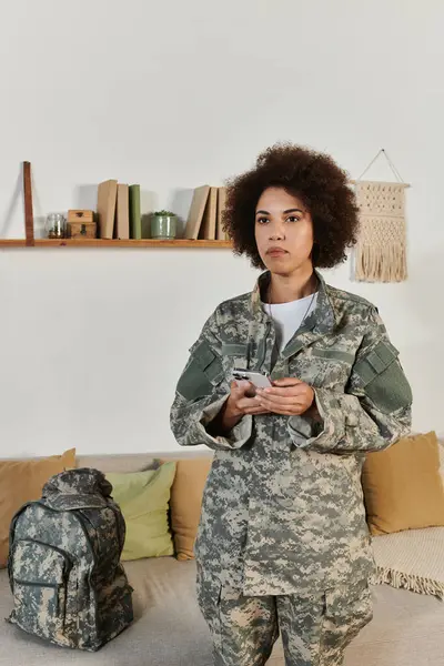 Una joven con equipo militar se para en su sala de estar, sosteniendo su teléfono y contemplando su próximo servicio. — Stock Photo