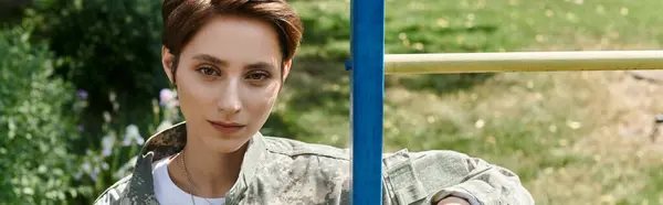 Bonita mujer soldado en uniforme de camuflaje de pie al aire libre, bandera - foto de stock
