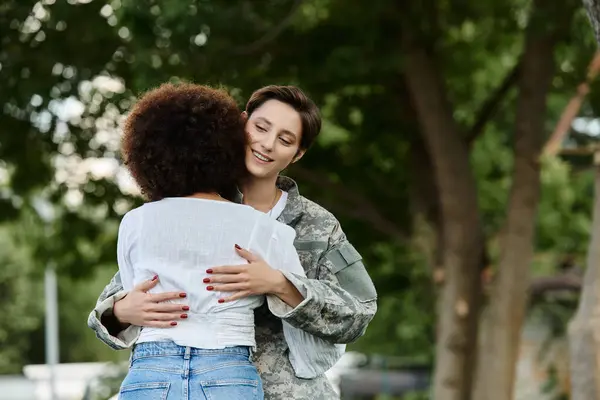 A female soldier in camouflage enjoys a heartfelt welcome from her wife in a picturesque park. — Stock Photo