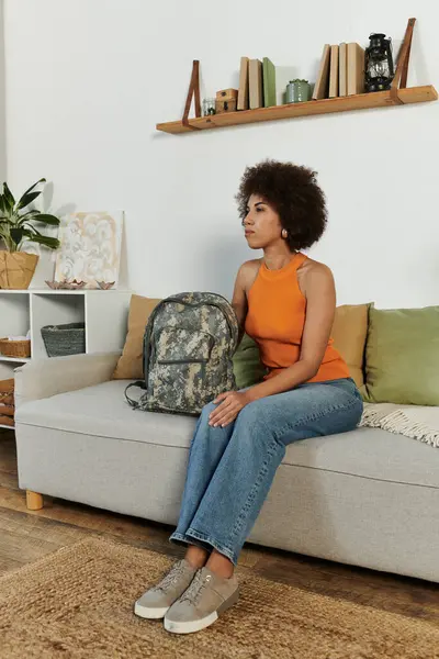 Una joven se sienta pensativamente en su sala de estar con una mochila de camuflaje a su lado. — Stock Photo