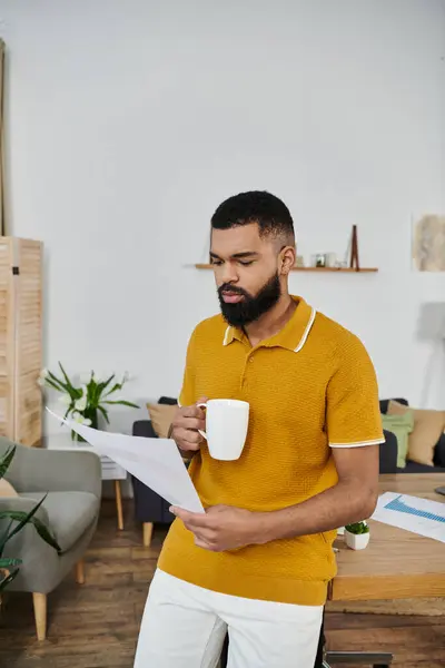 A man in a yellow sweater sips coffee while reviewing a document in his living space. — Stock Photo