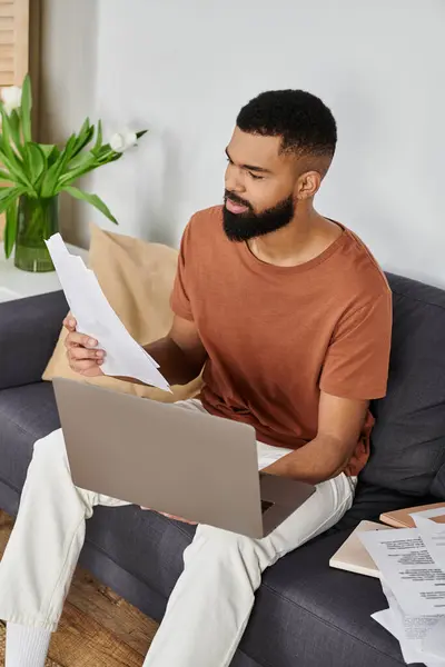 A man reviews documents while sitting on a couch with his laptop. — Stock Photo