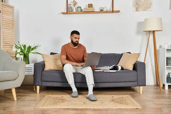 A man is seated comfortably on a sofa, working on his laptop with relaxed focus. — Stock Photo