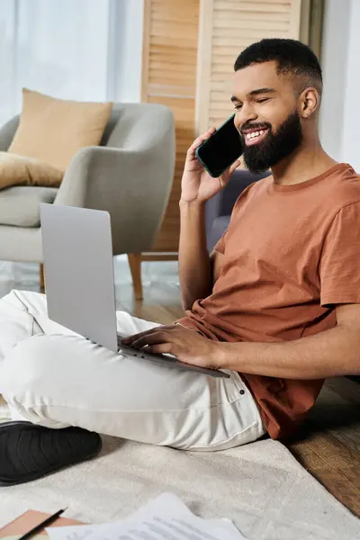A relaxed man smiles while balancing a phone call and laptop work at home. — Stock Photo