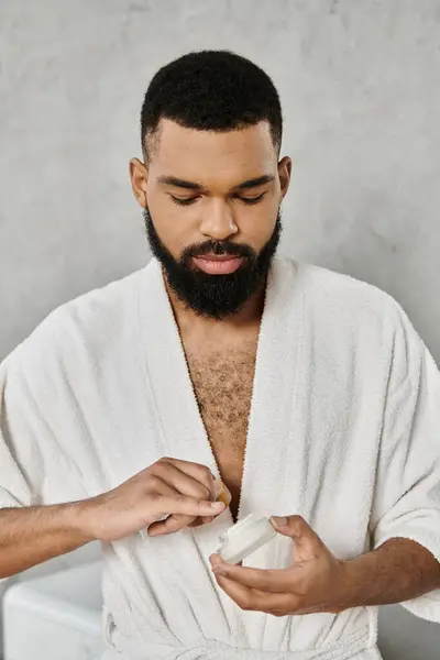 A man applies moisturizer while relaxing in a soft white robe. — Stock Photo