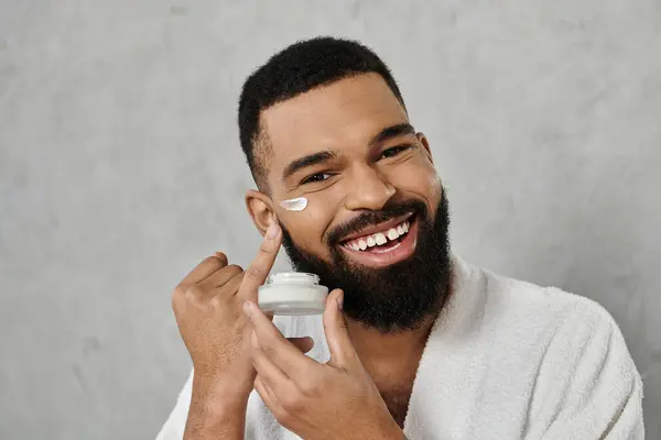 A cheerful man applies cream to his face while smiling at home. — Stock Photo