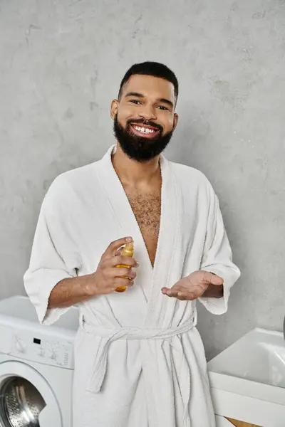 A relaxed man in a bathrobe smiles while holding a bottle at home. — Stock Photo