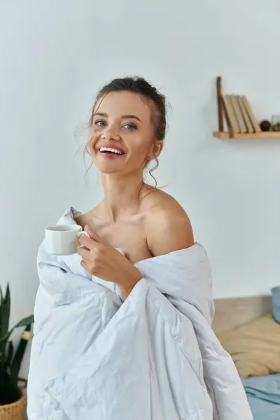 Una giovane donna avvolta in una coperta, sorridente con una tazza in mano. — Stock Photo