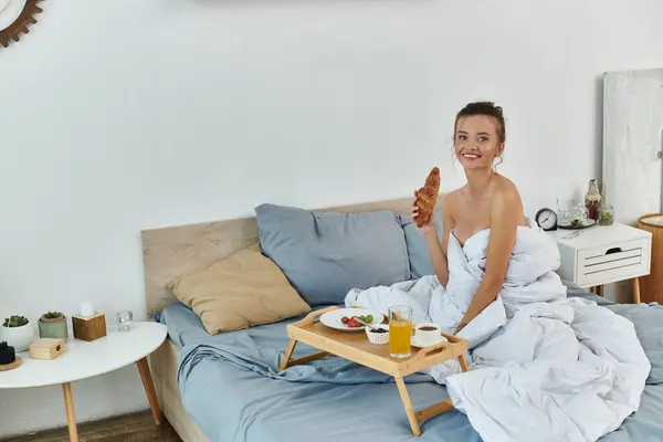 A young woman relaxes in bed, savoring breakfast while wrapped in sheets. — Stock Photo