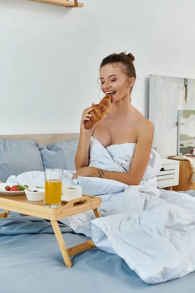 Uma jovem mulher saboreia um croissant e suco enquanto envolto em roupa de cama. — Fotografia de Stock