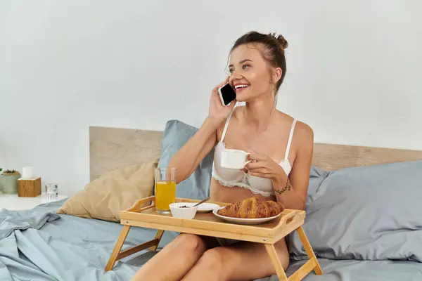 Cheerful young woman savoring breakfast in bed and chatting on her mobile phone. — Stock Photo