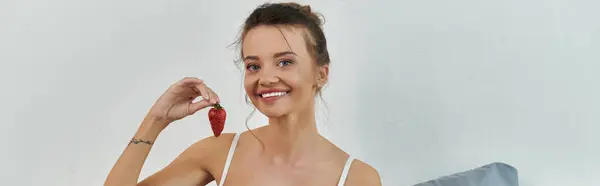 A joyful young woman playfully holds a strawberry while wearing elegant lingerie. — Stock Photo