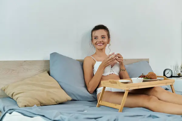 Une charmante jeune femme prend son petit déjeuner au lit, rayonnant de joie et de chaleur. — Photo de stock