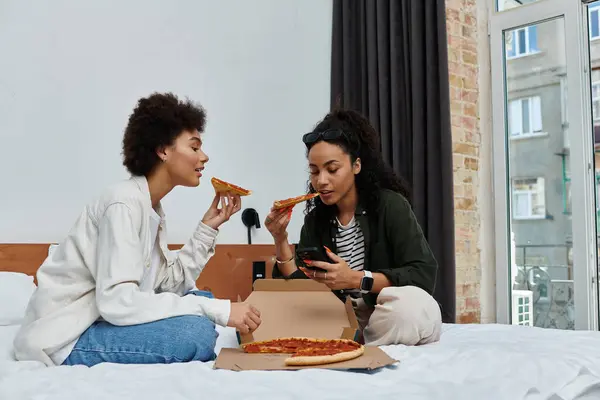 Zwei glückliche Frauen erfreuen sich an Pizza und Lachen, während sie sich in ihrem Hotelzimmer entspannen. — Stockfoto