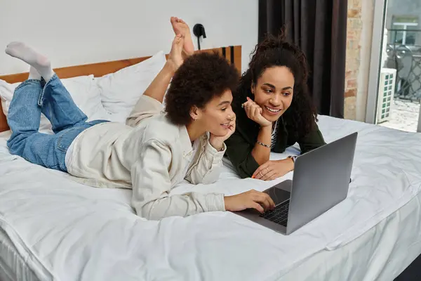 Deux femmes heureuses se détendent sur un lit, partageant des sourires tout en naviguant sur un ordinateur portable pendant leur voyage. — Photo de stock