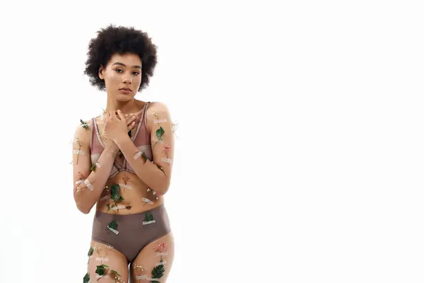 Délicieuse femme afro-américaine aux cheveux bouclés en sous-vêtements confortables pastel avec des fleurs sur le corps. — Photo de stock
