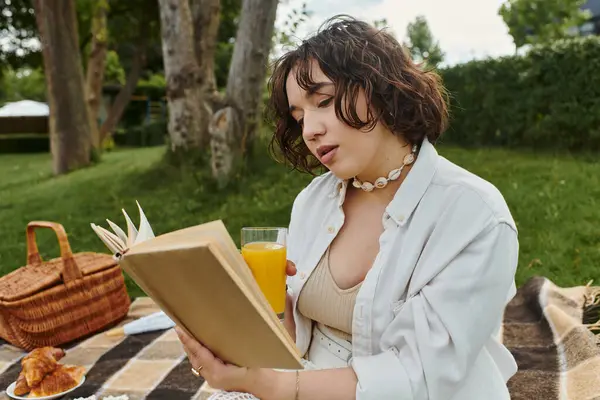 Una joven con una camisa blanca disfruta leyendo mientras toma una copa durante un picnic soleado de verano.. - foto de stock