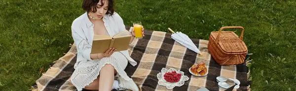 A young woman relaxes on a picnic blanket, sipping juice and immersed in a book under the summer sun. — Stock Photo