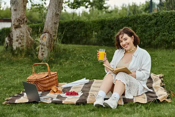 A cheerful woman relaxes on a blanket, savoring a refreshing drink while reading outdoors. — Stock Photo