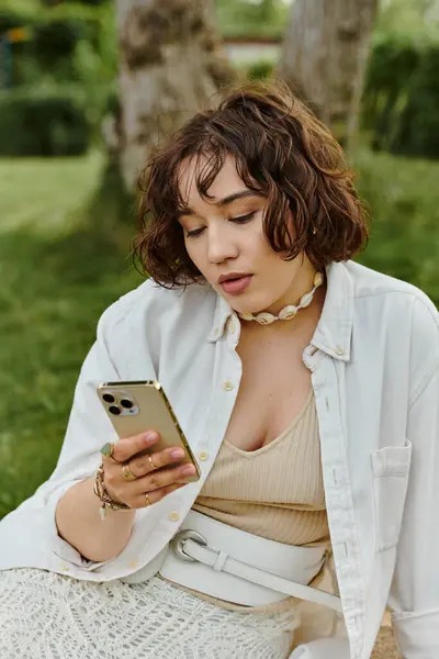 A pretty young woman takes a moment to relax during a summer picnic, engaged with her phone amidst the greenery. — Stock Photo