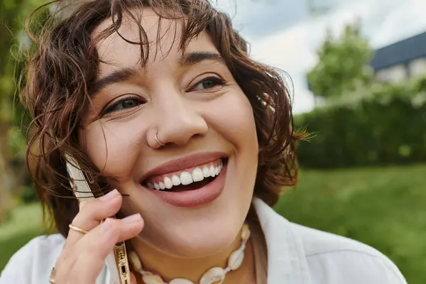 A cheerful young woman in a white shirt relaxes in the summer sun, chatting happily on her phone — Stock Photo