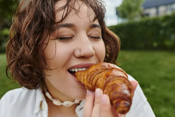 A joyful woman in a white shirt savors a flaky croissant under the warm summer sun. — Stock Photo