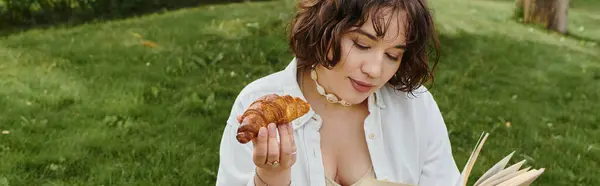 A young woman in a white shirt relaxes on a summer day, savoring a croissant while reading a book. — Stock Photo