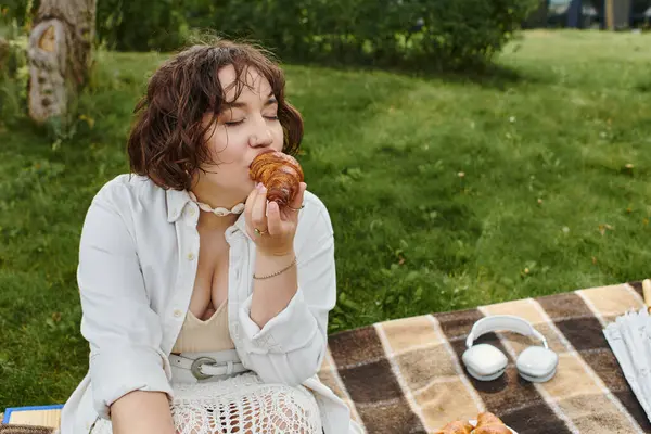 Una giovane donna in camicia bianca si rilassa su una coperta da picnic, deliziandosi in un croissant sotto il sole.. — Foto stock