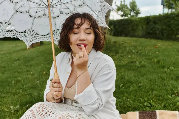 A lovely young woman enjoys a serene moment, applying lip balm under her delicate parasol in the summer sun. — Stock Photo