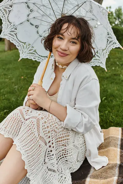 Uma jovem alegre relaxa em um cobertor, saboreando um piquenique de verão ensolarado com um guarda-chuva delicado. — Fotografia de Stock