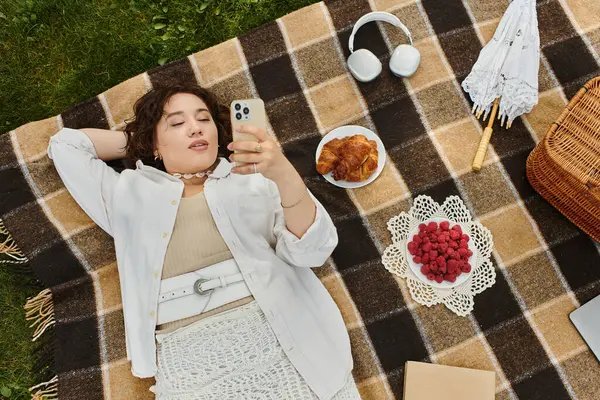 A young woman relaxes on a picnic blanket, savoring summer vibes while browsing her phone. — Stock Photo