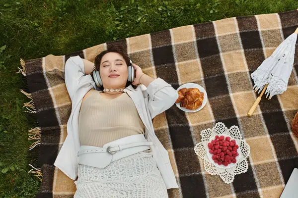 A young woman in a white shirt relaxes on a picnic blanket while enjoying music and tasty treats. — Stock Photo