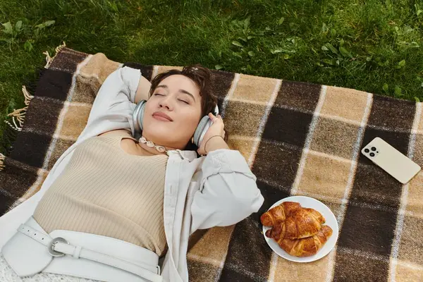 A young woman relaxes on a blanket, wearing headphones, while savoring croissants on a sunny day. — Stock Photo