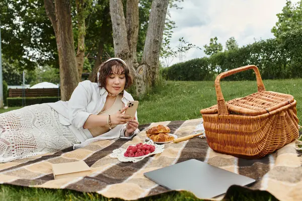 A young woman enjoys a sunny summer day, relaxing on a blanket with snacks while using her phone. — Stock Photo