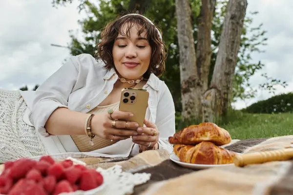 A young woman in a white shirt enjoys a summer picnic, relaxing with her phone and delicious pastries. — Stock Photo