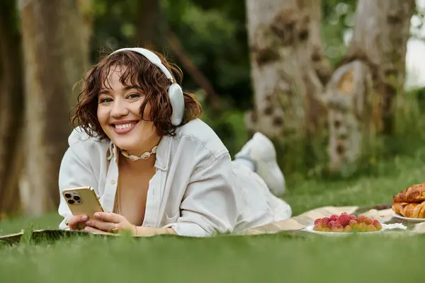 Una joven disfruta de un soleado día de verano, descansando sobre una manta de picnic con bocadillos y auriculares. - foto de stock