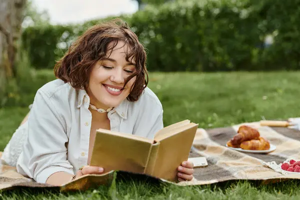 Uma jovem relaxa ao ar livre, imersa em um livro enquanto rodeada por um delicioso piquenique. — Fotografia de Stock