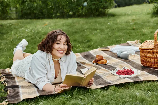A young woman enjoys a sunny summer day, reading a book while lounging on a picnic blanket filled with treats. — Stock Photo