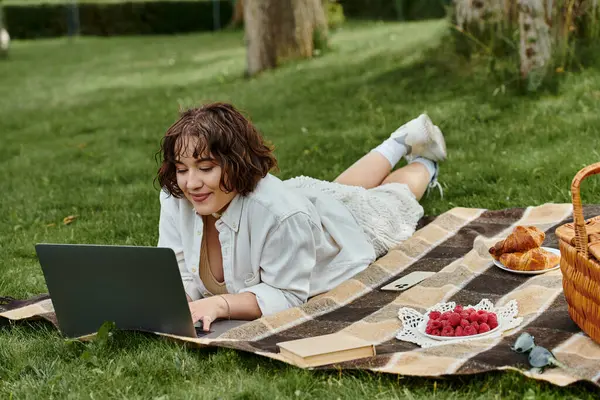 A young woman lounges on a picnic blanket, enjoying the summer sun while working on her laptop. — Stock Photo