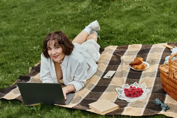A pretty young woman in a white shirt lounges on a blanket, enjoying a summer picnic with snacks and a laptop. — Stock Photo