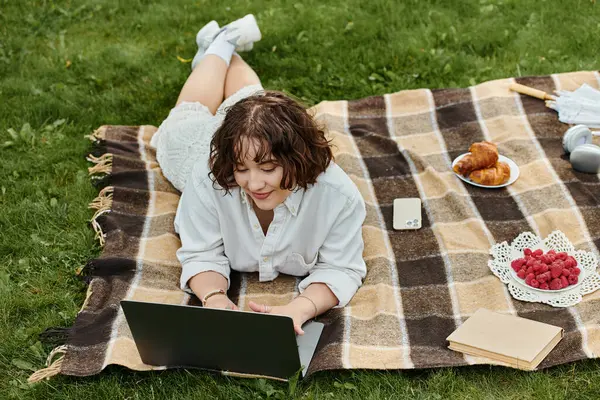 A young woman in a white shirt relaxes on a picnic blanket, working on her laptop amidst summer bliss. — Stock Photo