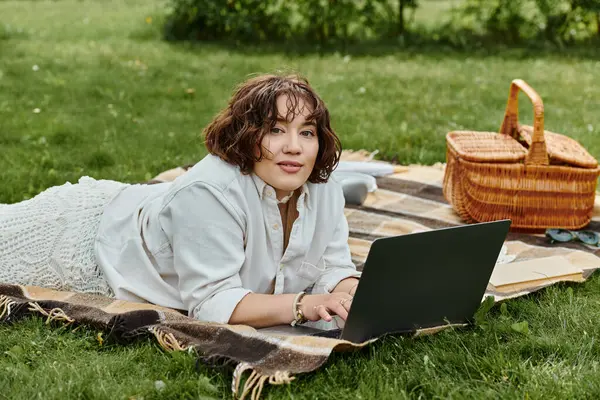 Eine junge Frau entspannt sich auf einer Picknickdecke, genießt die Sonne und arbeitet im Sommer an ihrem Laptop. — Stockfoto