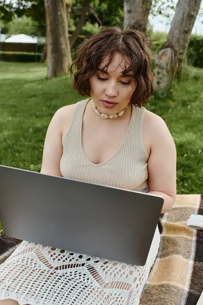 Uma jovem mulher em uma camisa branca relaxa em um cobertor de piquenique, absorvido em seu laptop em meio a um dia ensolarado de verão. — Stock Photo