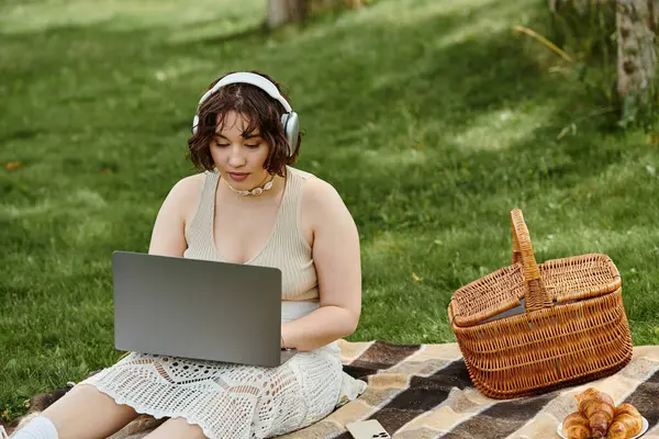 A young woman in a white shirt relaxes on a picnic blanket, focused on her laptop amid summer greenery. — Stock Photo