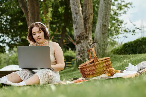 A young woman relaxes during a summer picnic, listening to music while working on her laptop in the park. — Stock Photo